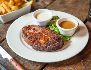 A plated steak, 2 accompanying sauce cups, and a plate of fries sitting on a table