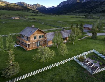 Light brown cabin with two stories and a light gray roof in the middle of a field surrounded by mountains.