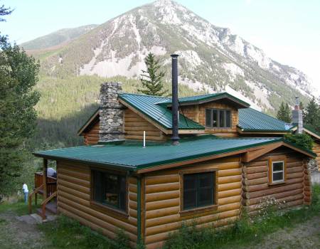Brown cabin with a teal roof surrounded by trees, in front of a forested mountain.