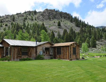 Brown one-story cabin in the middle of a clear field, in front of a forested hill.