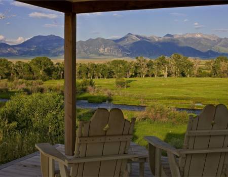 Two wooden chairs on a back patio looking out at a field, forest, and a range of mountains.