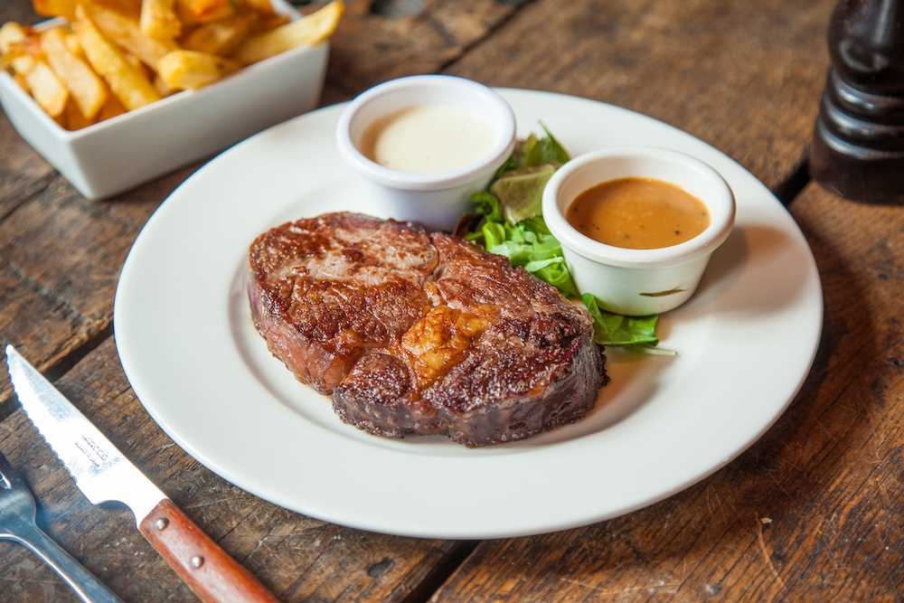 A plated steak, 2 accompanying sauce cups, and a plate of fries sitting on a table